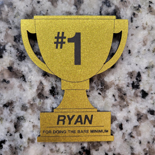 Gold trophy-shaped card with '#1 NAME FOR DOING THE BARE MINIMUM' text on a granite surface