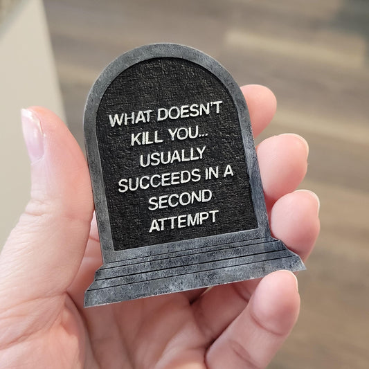 Small tombstone-shaped sign with humorous text held in a hand against a blurred background