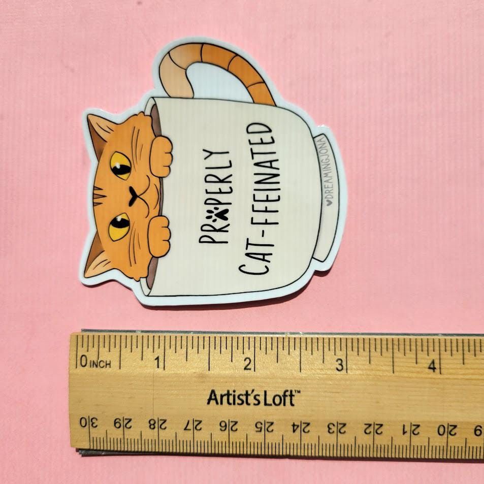 Sticker of a cat peeking out from a coffee cup with text, next to a ruler for size reference on a pink background.
