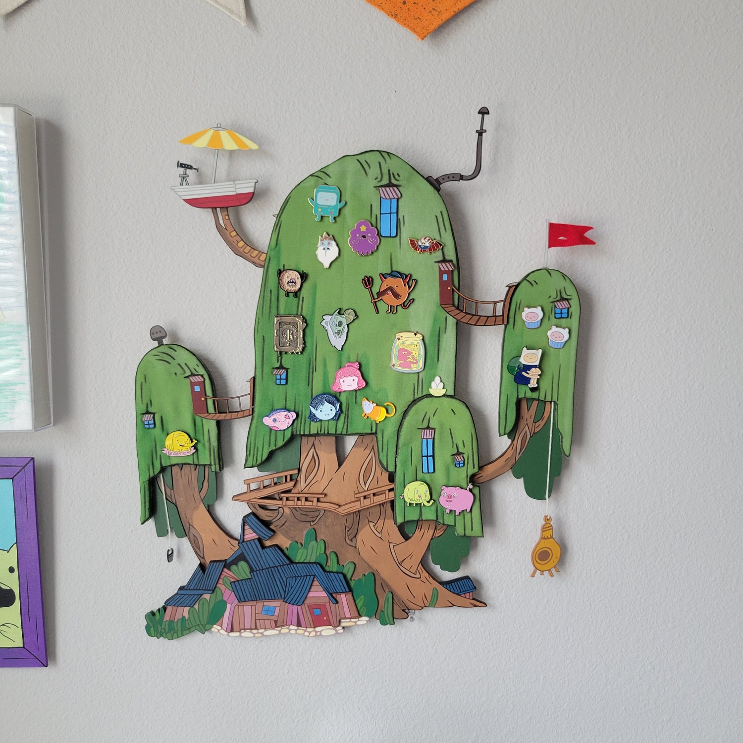 Colorful wall art of the Adventure Time treehouse with various small enamel pins tacked on.