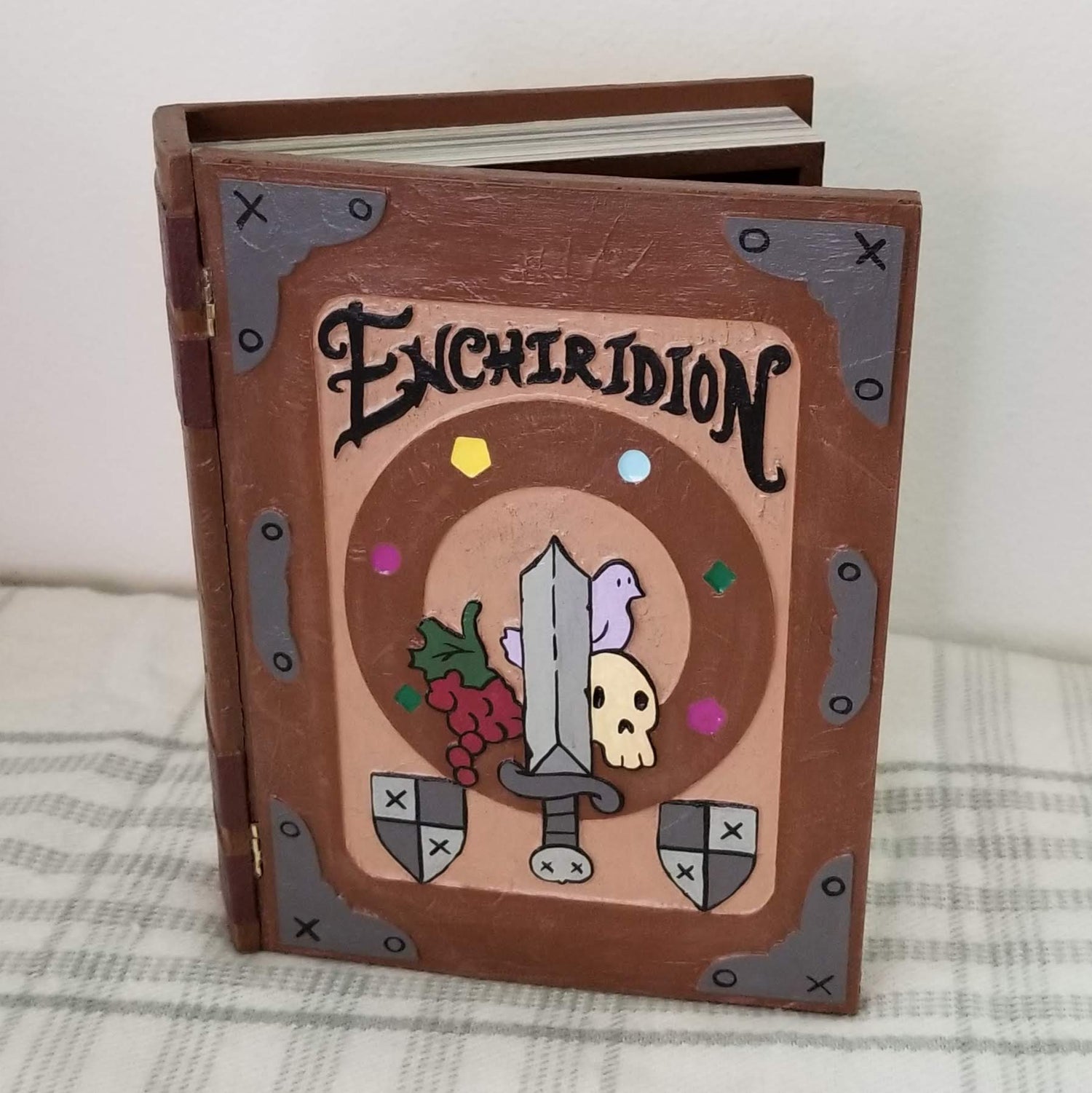 Decorative book titled 'Enchiridion' with a sword and skull design on a checkered fabric background