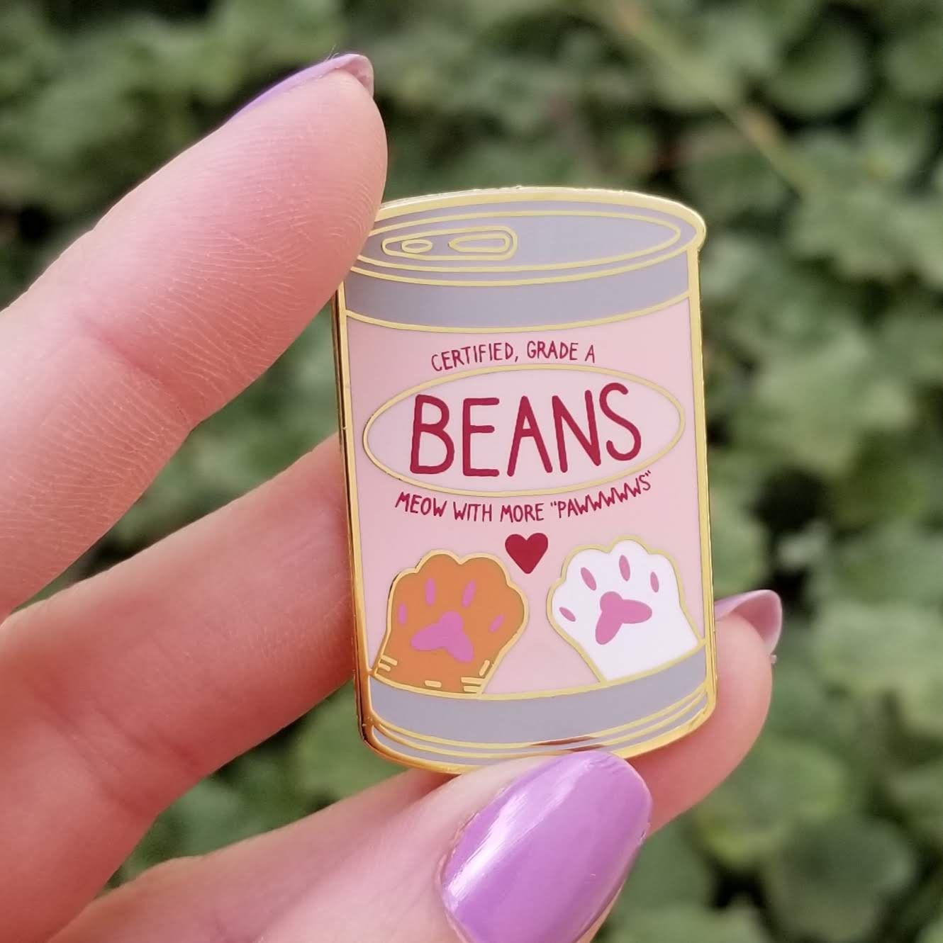 Enamel pin of a can labeled 'Beans' held by a hand with purple nail polish against a green leafy background.