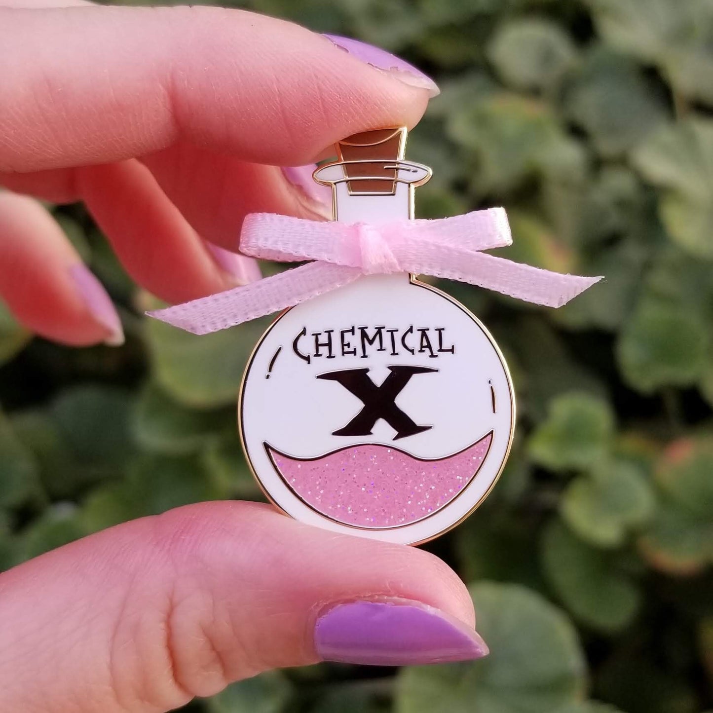 Enamel pin shaped like a bottle labeled 'Chemical X' held by a hand with purple nail polish against a green leafy background.