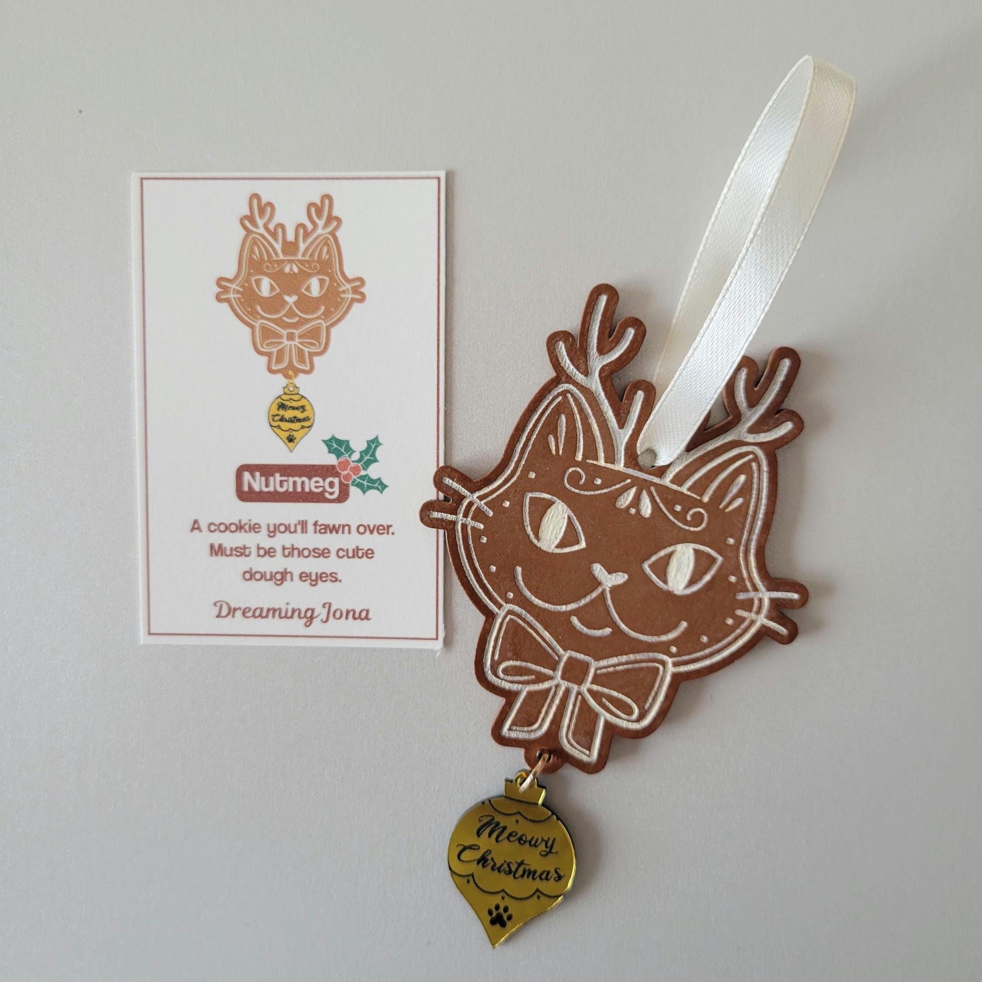 Cat-shaped ornament with a cream ribbon and 'Meowy Christmas' gold tag on a white background