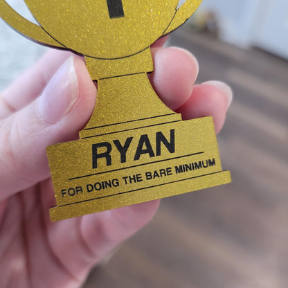 Gold trophy-shaped award with 'NAME FOR DOING THE BARE MINIMUM' text held by a hand.