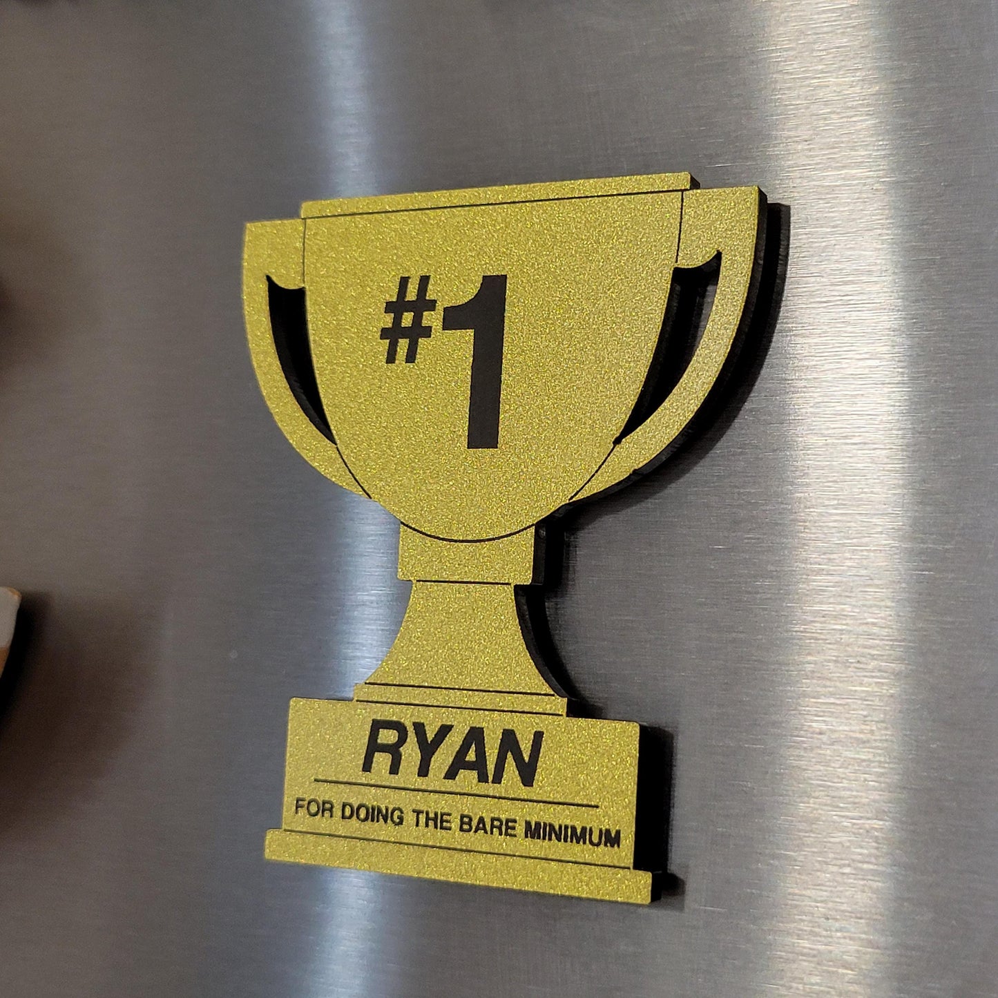 Gold trophy-shaped magnet with '#1 NAME FOR DOING THE BARE MINIMUM' text on a fridge