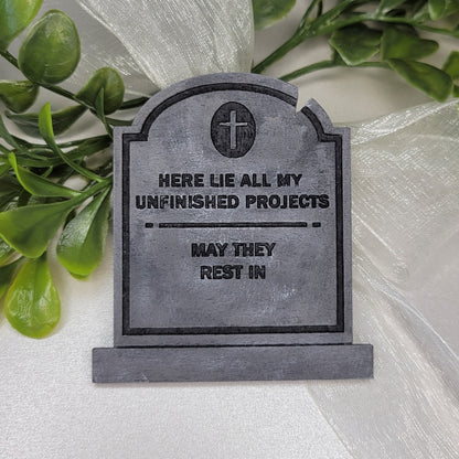 Wooden painted tombstone with words 'here lie all my unfinished projects may they rest in'