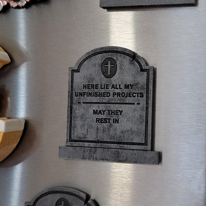 Fridge with a tombstone-shaped wooden magnet reading 'Here lie all my unfinished projects, may they rest in.'