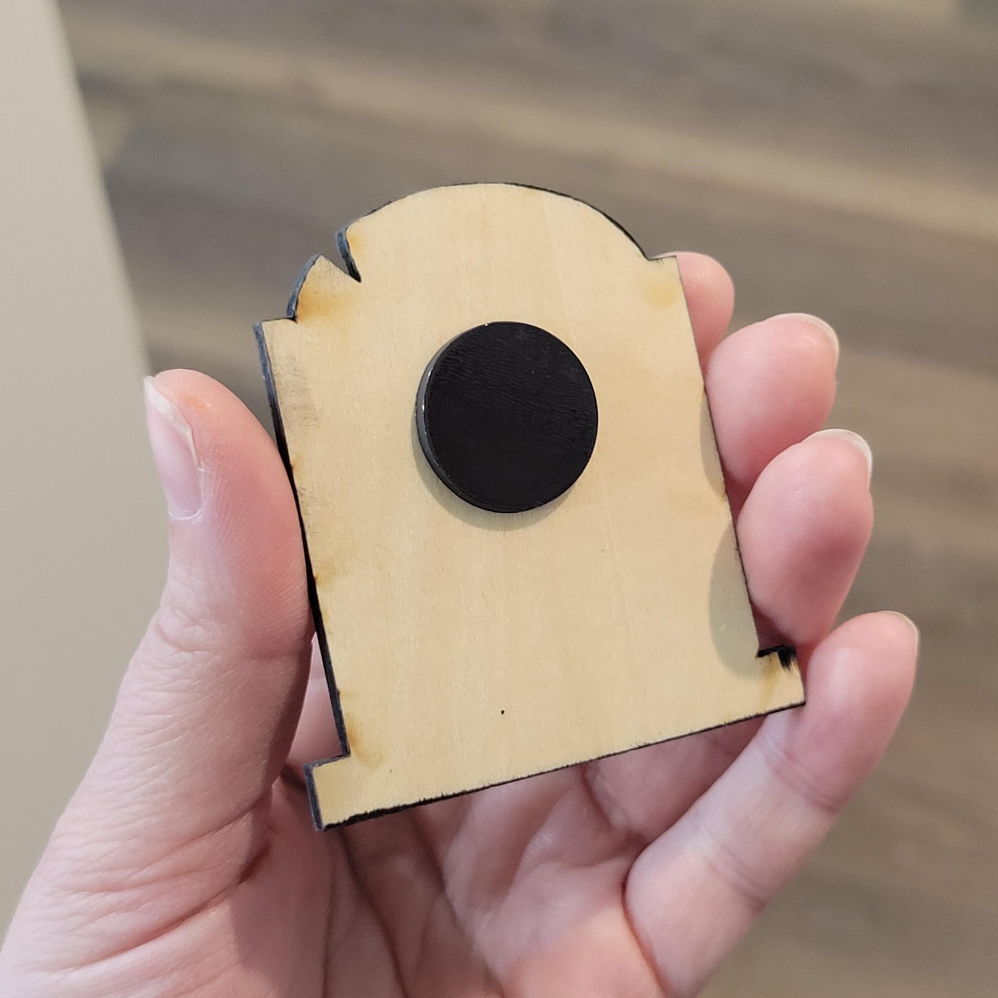 Hand holding a small wooden tombstone with a black magnet against a blurred background