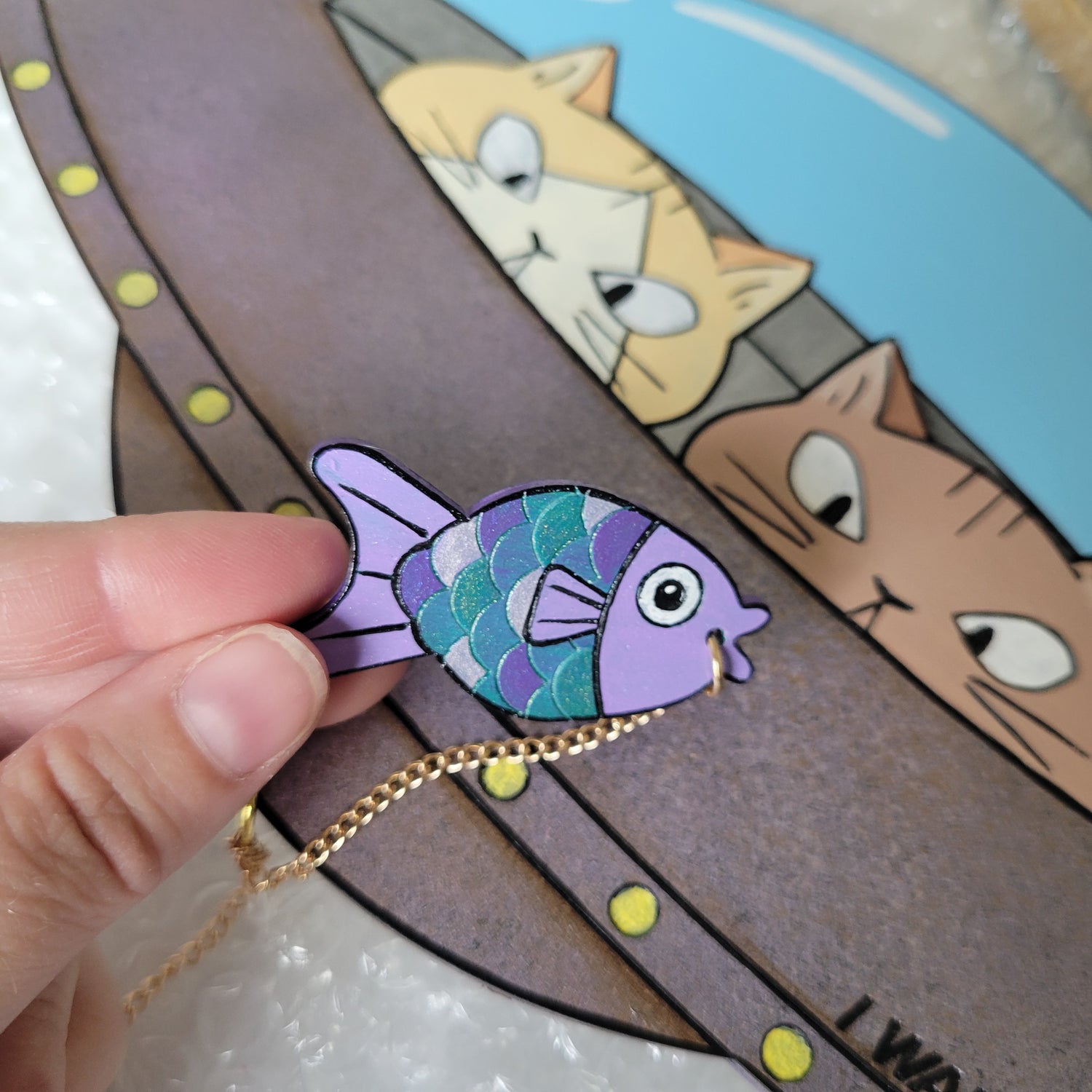 Hand holding a colorful fish-shaped keychain with cartoon cat faces in the background in a UFO