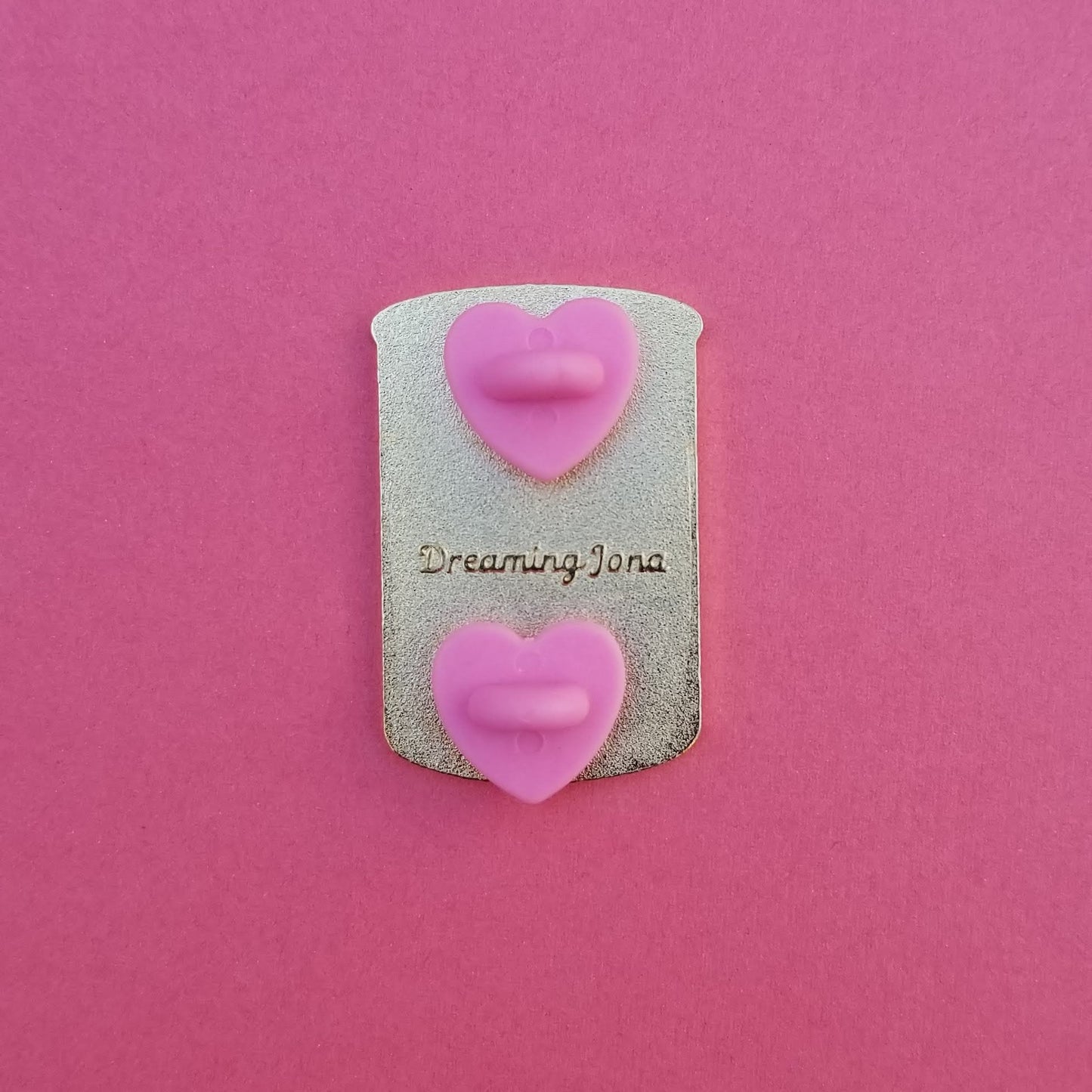 Decorative pin with pink backing posts and 'Dreaming Jona' text on a pink background