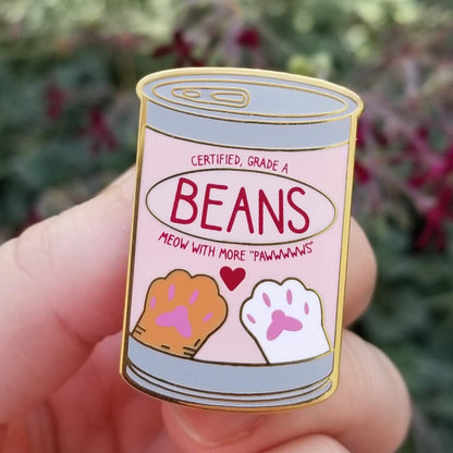 Enamel pin shaped like a can of 'Beans' with paw prints and text held in a hand against a blurred floral background.