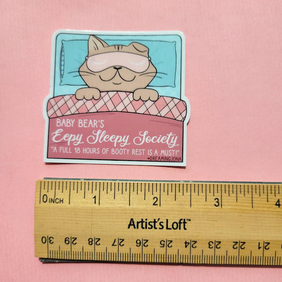 Sticker of a bear in pajamas with a sleep mask on a pink background, next to a ruler for scale.
