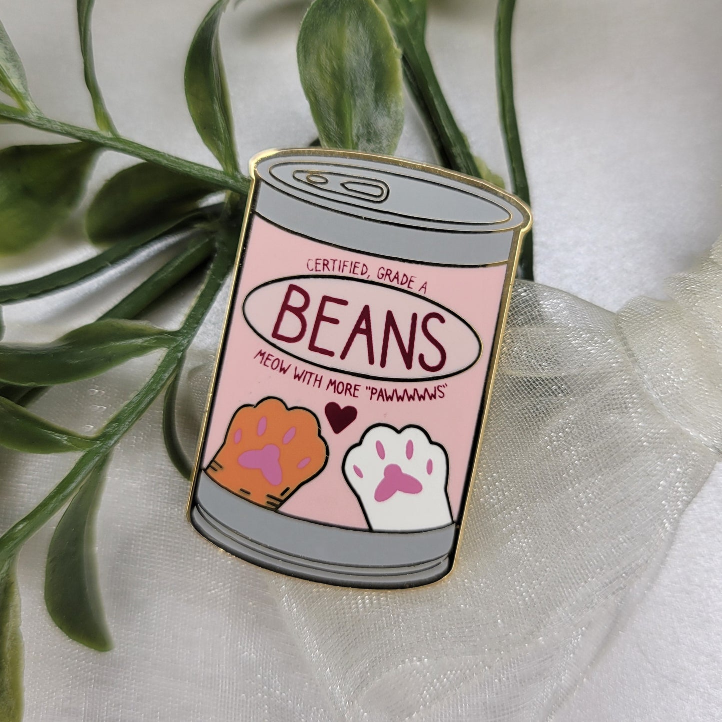 Enamel pin shaped like a can labeled 'Beans' on a white background with green leaves.