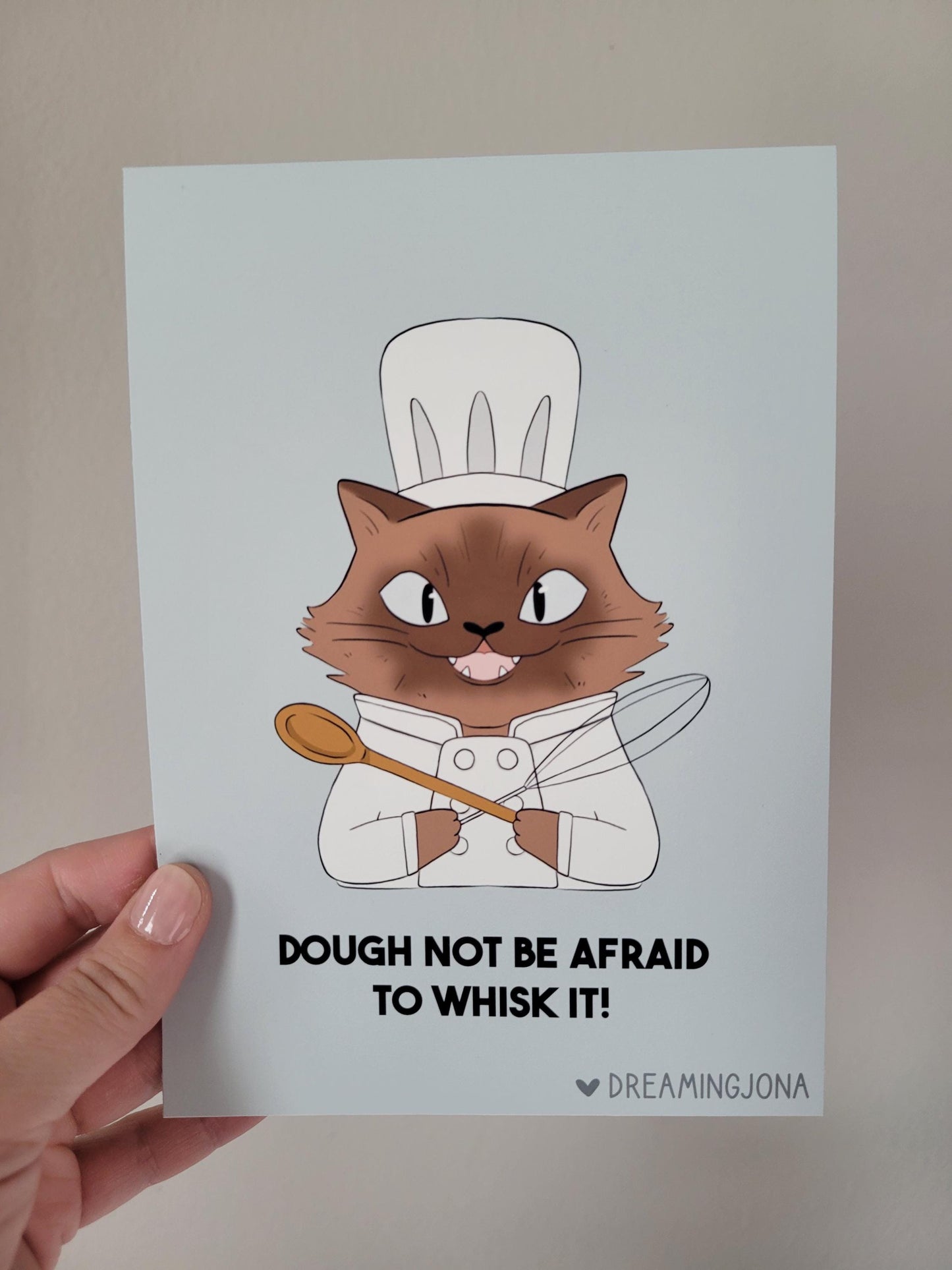 Card with a cat chef illustration and text 'Dough not be afraid to whisk it!' held by a hand.
