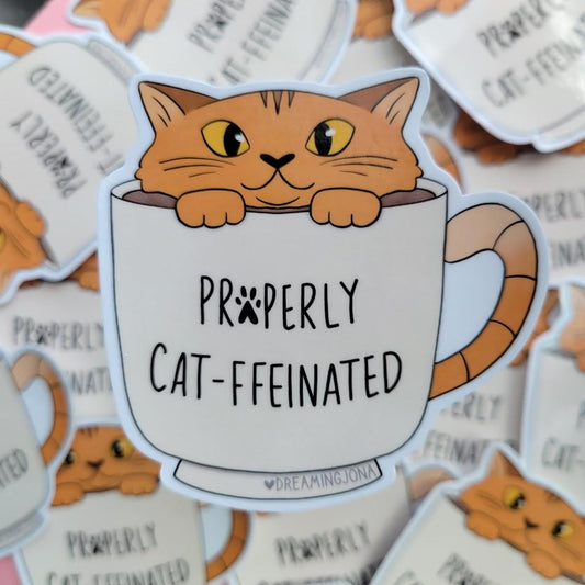 Sticker of a cat in a coffee cup with 'Properly Cat-ffeinated' text on a background of similar stickers.