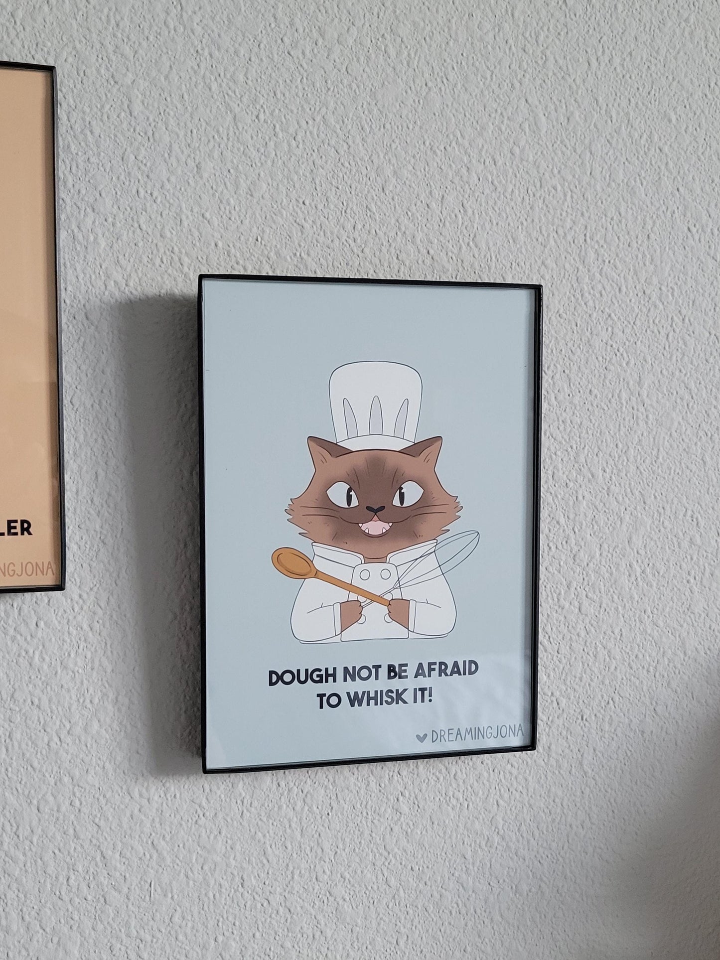 Framed poster of a cat chef holding a wooden spoon with text on a light gray wall.