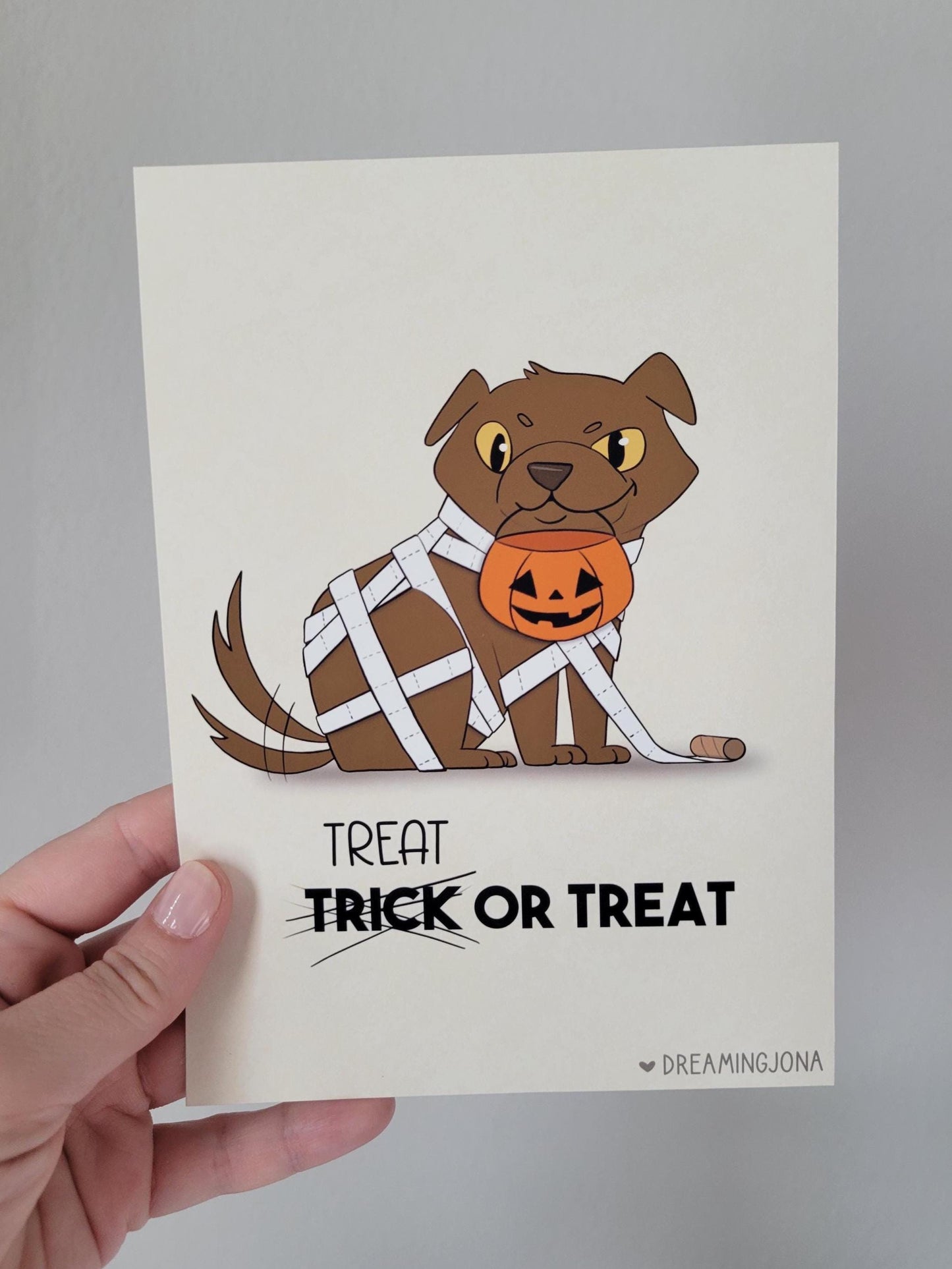 Halloween-themed card with a dog wearing a pumpkin costume, held by a hand against a gray background.