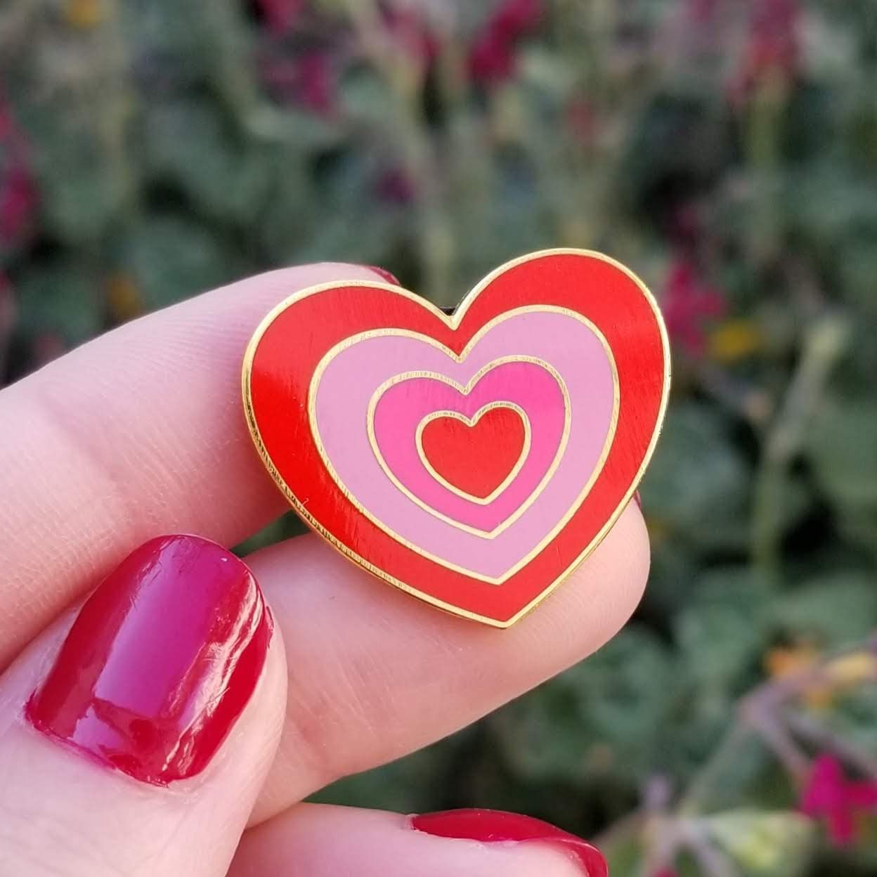 Heart-shaped pin with red and pink hearts held between fingers against a blurred natural background