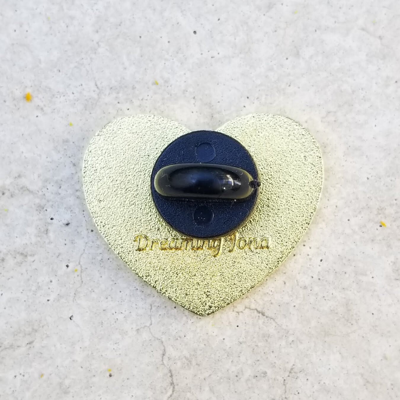 Heart-shaped pin with a backing post and 'Dreaming John' text on a textured surface
