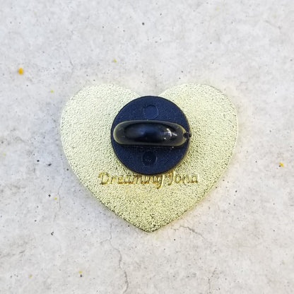 Heart-shaped pin with a backing post and 'Dreaming John' text on a textured surface