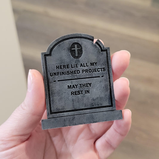 Small tombstone-shaped object with humorous text held in a hand against a blurred background