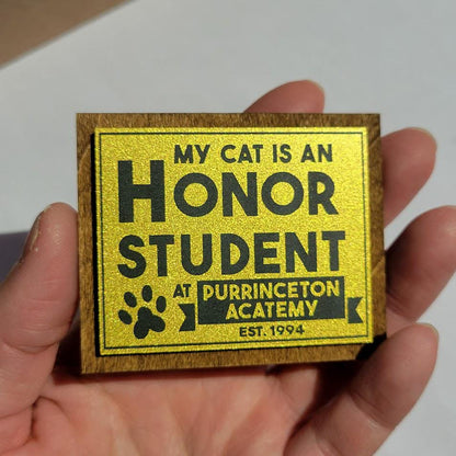 gold and black sign with text 'My cat is an honor student at Purrinceton Academy' held in a hand.