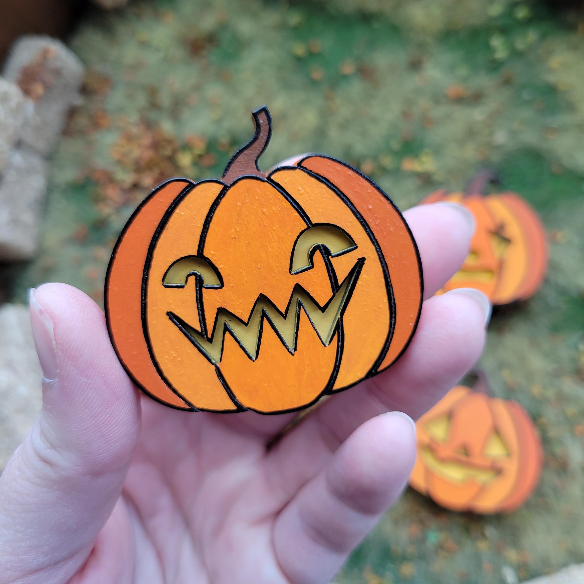 Hand holding a paper pumpkin cutout with a carved face against a blurred natural background