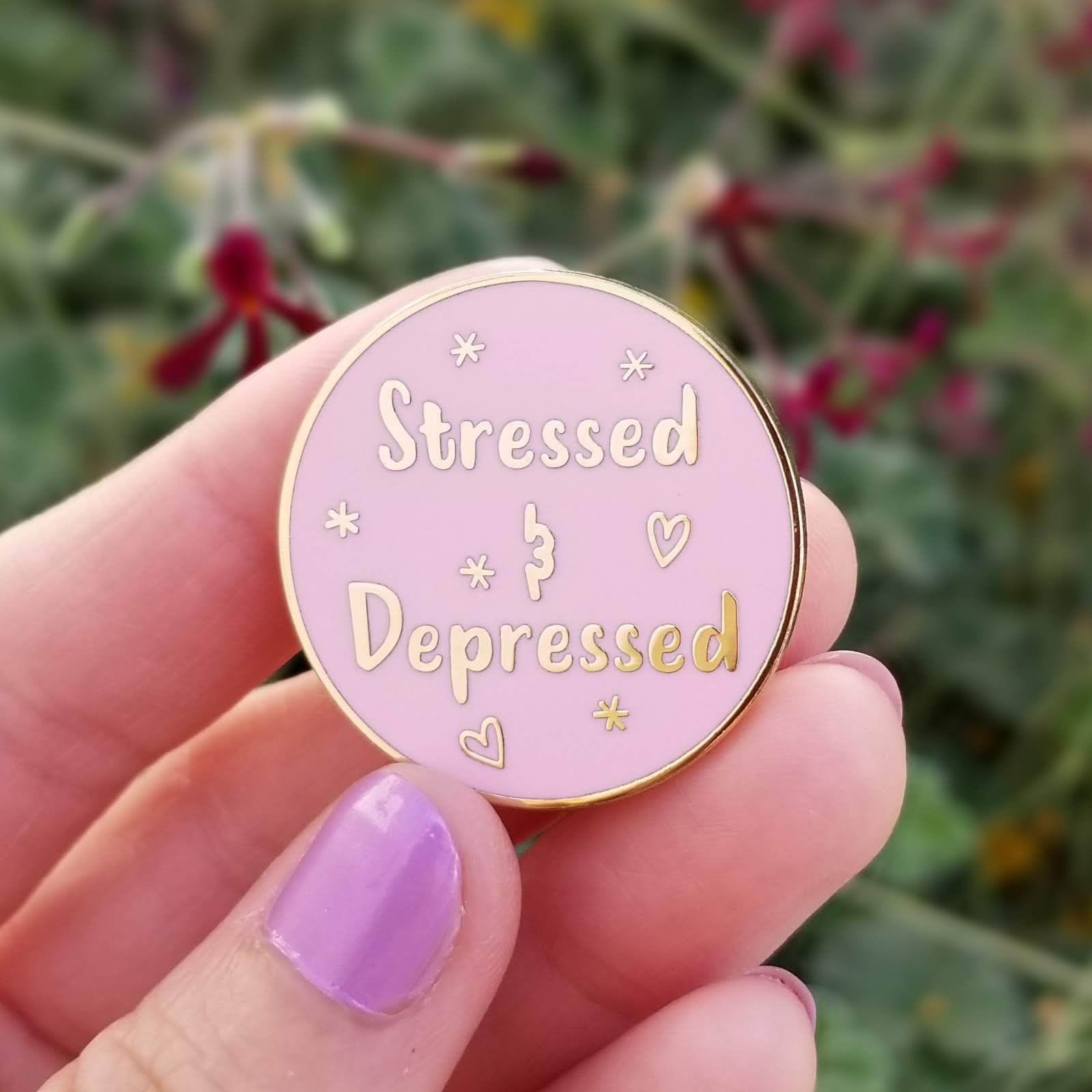Pink enamel pin with 'Stressed & Depressed' text held by a hand with purple nail polish against a blurred natural background.
