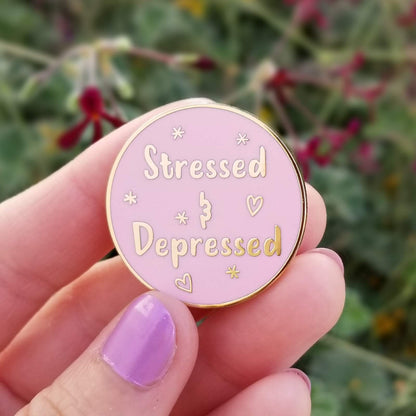 Pink enamel pin with 'Stressed & Depressed' text held by a hand with purple nail polish against a blurred natural background.