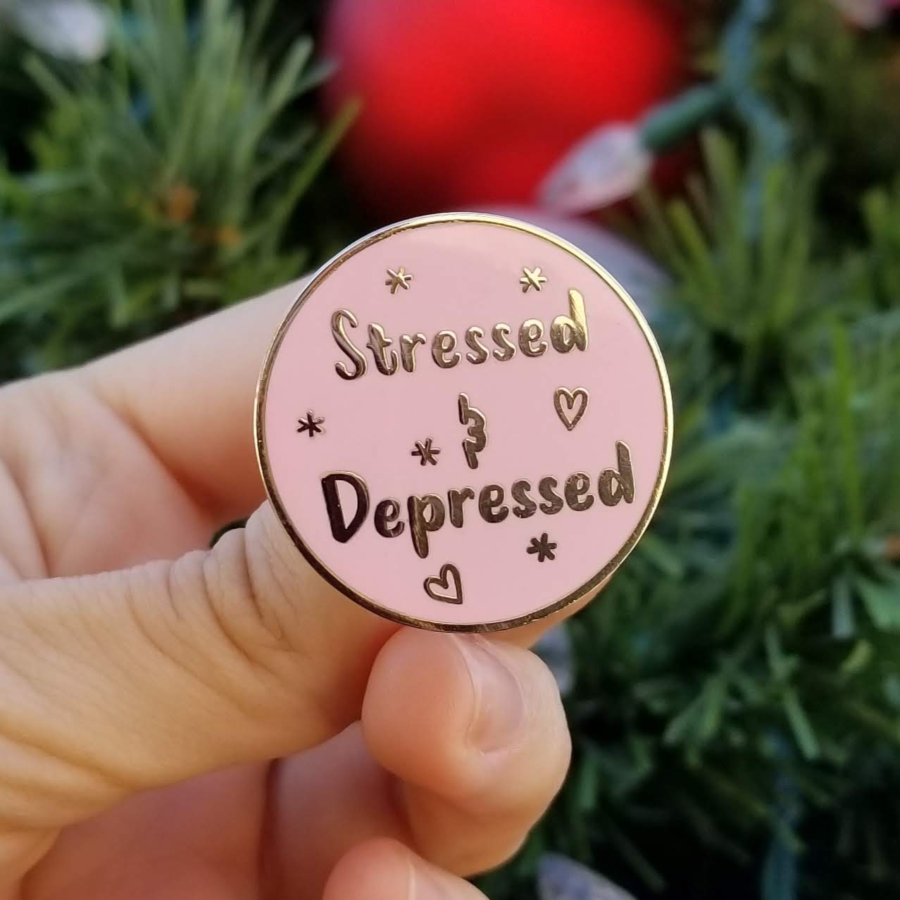 Pink enamel pin with 'Stressed & Depressed' text held by a hand against a blurred green and red background.