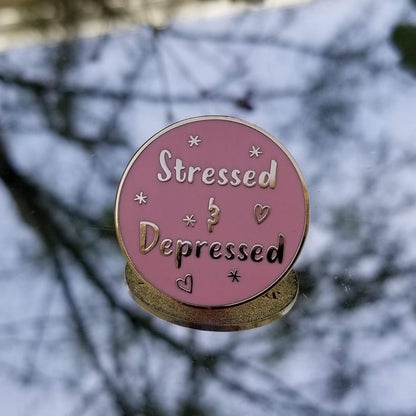 Pink enamel pin with 'Stressed & Depressed' text against a blurred mirror background
